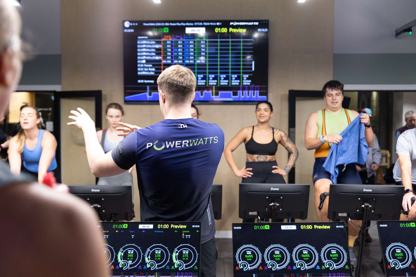 A PowerWatts coach leading a session, riders and live power data screens behind
